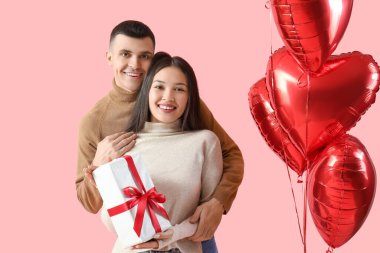 Sevgililer Günü hediyesi olan genç bir çift ve pembe arka planda kalp şeklinde balonlar.