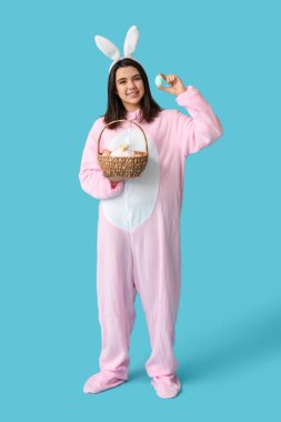 Tavşan kostümlü güzel bir kadın elinde hasır sepetle Paskalya pastası ve mavi arka planda yumurtalar tutuyor.