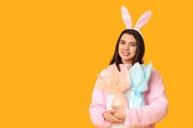 Sarı arka planda Paskalya hediyesi yumurtaları olan tavşan kostümlü mutlu genç bir kadın.