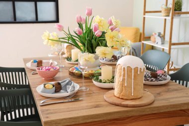 Şenlikli masa dekorları, boyanmış yumurtalar, paskalya pastası ve vazo dolusu çiçekler yemek odasında.