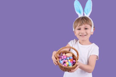 Tavşan kulaklı şirin bir çocuk elinde sepetle mor arka planda Paskalya yumurtaları tutuyor.