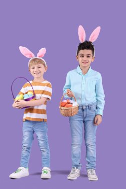 Tavşan kulaklı şirin çocuklar mor arka planda paskalya yumurtaları taşıyan hasır sepetler tutuyorlar.