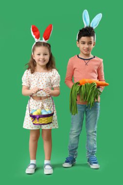Tavşan kulaklı şirin çocuklar ellerinde hasır sepet ellerinde paskalya yumurtaları ve yeşil arka planda oyuncak havuç.