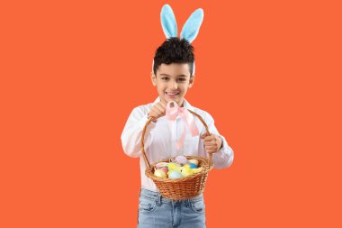 Tavşan kulaklı şirin çocuk elinde hasır sepetle turuncu arka planda Paskalya yumurtaları tutuyor.