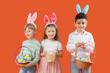 Tavşan kulaklı şirin çocuklar ellerinde hasır sepetlerle turuncu arka planda Paskalya yumurtaları taşıyorlar.