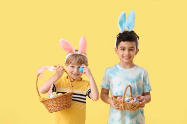 Tavşan kulaklı şirin çocuklar ellerinde sepetler ve sarı arka planda Paskalya yumurtaları var.