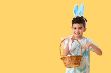 Tavşan kulaklı şirin çocuk sarı arka planda paskalya yumurtaları olan sepeti işaret ediyor.
