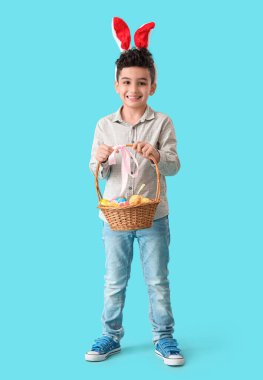 Tavşan kulaklı şirin çocuk elinde sepet, mavi arka planda Paskalya yumurtaları tutuyor.