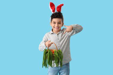 Tavşan kulaklı şirin çocuk hasır sepeti işaret ediyor mavi arka planda oyuncak havuçları var. Paskalya kutlaması