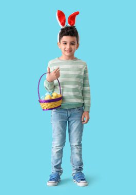 Tavşan kulaklı şirin çocuk elinde sepet, mavi arka planda Paskalya yumurtaları tutuyor.