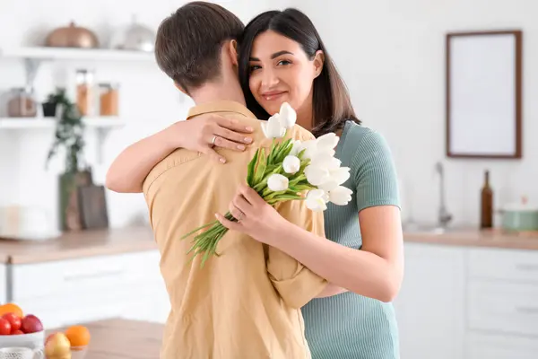 Young pregnant woman with tulips hugging her husband in kitchen
