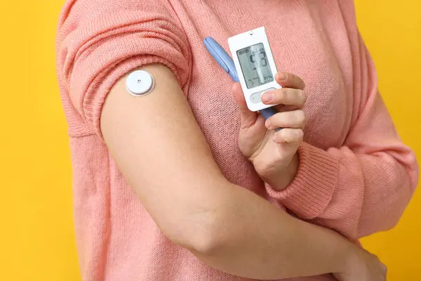 stock image Woman with glucometer, lancet pen and sensor for measuring blood sugar level on yellow background, closeup. Diabetes concept