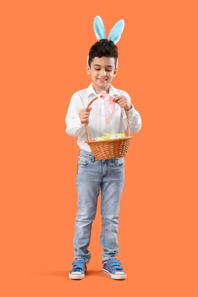 オレンジ色の背景にイースターエッグとウィッカーバスケットを保持するバニー耳のかわいい男の子 — ストック写真