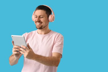 Mavi arka planda tablet bilgisayar kullanan kulaklıklı yakışıklı bir adam.