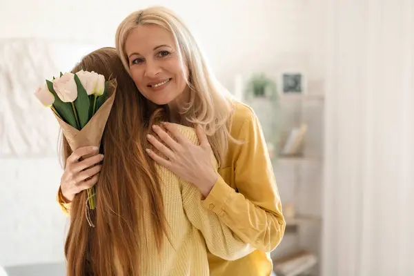 Mature woman with tulips hugging her daughter at home. Mother\'s Day celebration