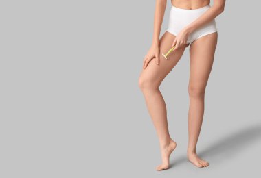 Beautiful young woman with razor shaving legs on grey background, closeup