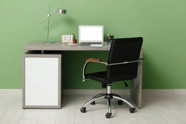 Yeşil duvarın yanında dizüstü bilgisayarı, lambası ve koltuğu olan modern ofis işyeri