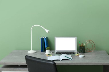 Yeşil duvarın yanında dizüstü bilgisayarı, kırtasiyesi ve lambası olan modern ofis işyeri