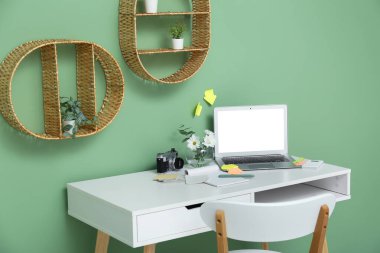 Yeşil duvarın yanında dizüstü bilgisayarı, çiçekleri, kırtasiyesi ve fotoğraf makinesi olan modern ofis işyeri