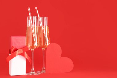 Kırmızı arka planda kağıt kalp ve hediye kutusu olan şampanya kadehleri. Sevgililer Günü kutlaması