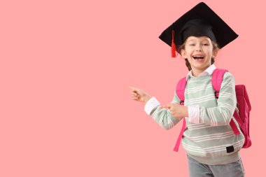 Mezuniyet şapkalı küçük kız pembe arka plandaki bir şeyi işaret ediyor. Okul yılı sonu.