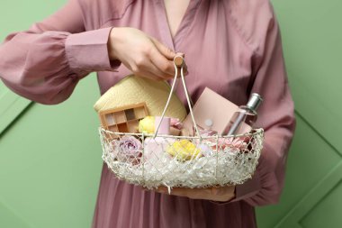 Yeşil arka planda dekoratif kozmetik ve çiçeklerle dolu sepeti olan güzel genç bir kadın.