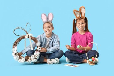Mavi arka planda oturan paskalya yumurtası ve çelengi olan tavşan kulaklı küçük çocuklar.