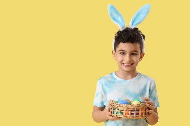 Tavşan kulaklı şirin bir çocuk elinde sepet ve sarı arka planda Paskalya yumurtaları tutuyor.