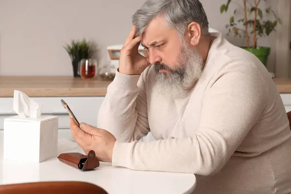 Portrait of sad senior man with smartphone sitting at table in kitchen