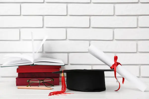 Mortar board with diploma, eyeglasses and books on table near white brick wall