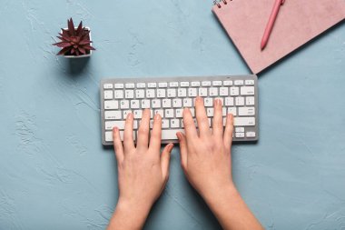 Mavi arkaplanda dizüstü bilgisayarın yanında bilgisayar klavyesiyle yazı yazan kadın elleri