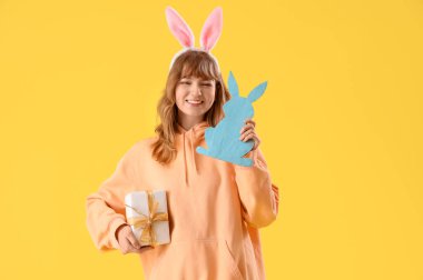 Paskalya tavşanı kulaklı genç kız, kağıttan tavşan ve sarı arka planda hediye.