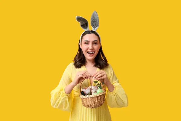 黄色の背景に装飾的な化粧品とイースターのバスケットを保持するバニーの耳の若い女性 — ストック写真