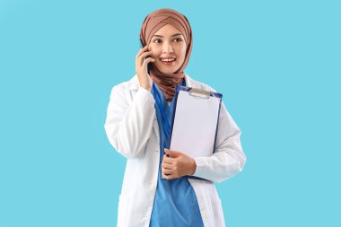 Not panosuyla konuşan kadın Müslüman doktor mavi arka planda cep telefonuyla konuşuyor