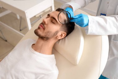 Klinikte genç bir adamın saçını inceleyen büyüteçli bir doktor.