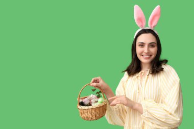 Tavşan kulaklı güzel bir kadın yeşil arka planda kozmetik ürünlerle dolu bir Paskalya sepeti tutuyor.