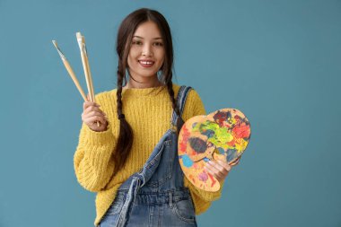 Mavi arka planda fırça ve boya paleti olan Asyalı bir kadın sanatçı.