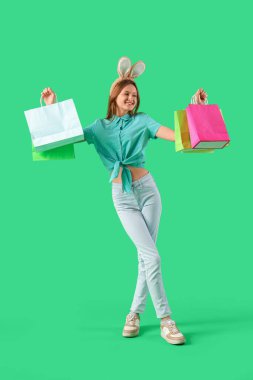 Yeşil arka planda alışveriş torbaları olan tavşan kulaklı genç bir kadın. Paskalya kutlaması