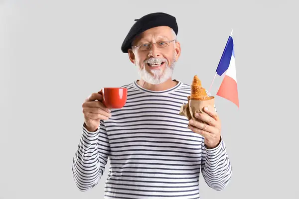 Senior man with flag of France, coffee cup and croissant on light background