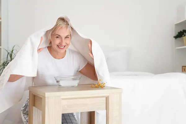 Mature woman doing steam inhalation at table in bedroom