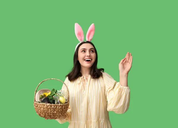 バニーの耳に幸せな若い女性は緑の背景に化粧品の製品とイースターのバスケットを保持しています — ストック写真