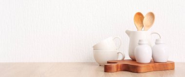 Beyaz duvarın yanındaki ahşap masada mutfak gereçleri ve çanak çömlek seti. Tasarım için pankart