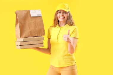 Kese kağıdı ve pizza kutuları olan kadın kurye sarı arka planda baş parmağını kaldırıyor.
