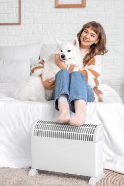 Sevimli Samoyed köpeği olan genç bir kadın yatak odasındaki radyatörde bacaklarını ısıtıyor.