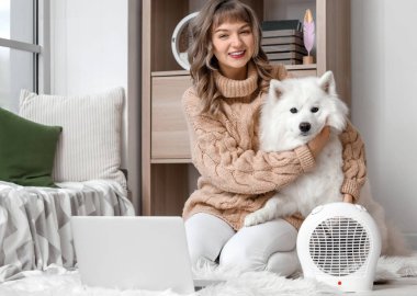 Evde elektrikli fan ısıtıcısının yanında Samoyed köpeği ve dizüstü bilgisayarı olan genç bir kadın.