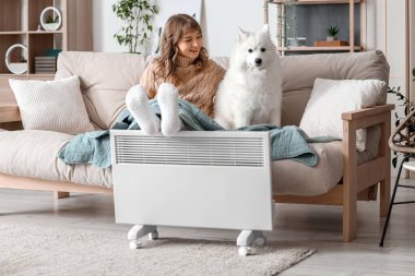 Ekoseli genç bir kadın ve Samoyed köpeği radyatörde bacaklarını ısıtıyorlar.