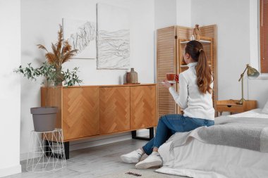 Elinde bir fincan kahveyle yatak odasındaki 3 boyutlu resimlere bakan genç bir kadın.