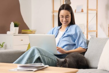 Evdeki kanepede dizüstü bilgisayarı ve sevimli İngiliz Shorthair kedisi olan genç bir kadın.