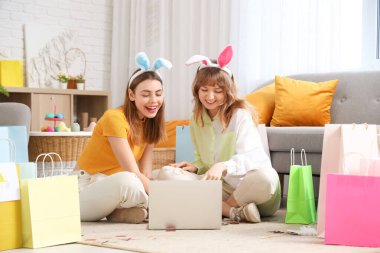 Paskalya 'da evde otururken dizüstü bilgisayarı ve kese kağıdı olan tavşan kulaklı mutlu genç kadınlar. İnternetten alışveriş