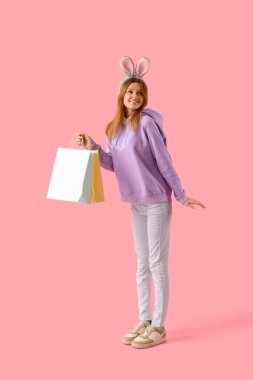 Pembe arka planda alışveriş torbaları olan tavşan kulaklı genç bir kadın. Paskalya kutlaması
